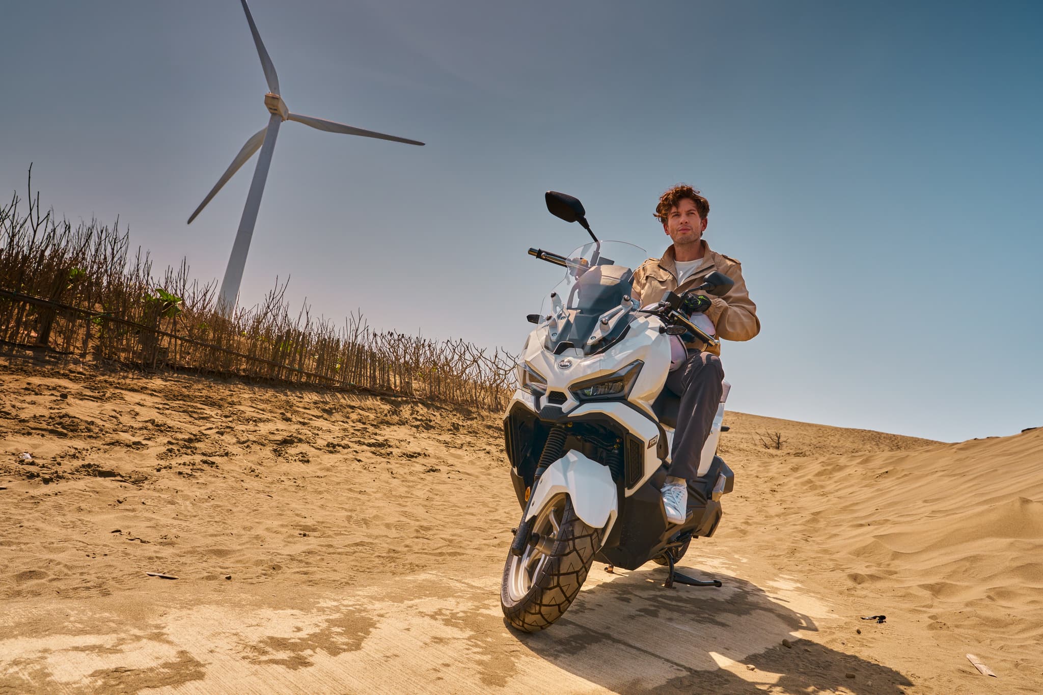 Hombre en scooter SYM ADX 125 ABS TCS blanca en un paisaje desértico con aerogenerador