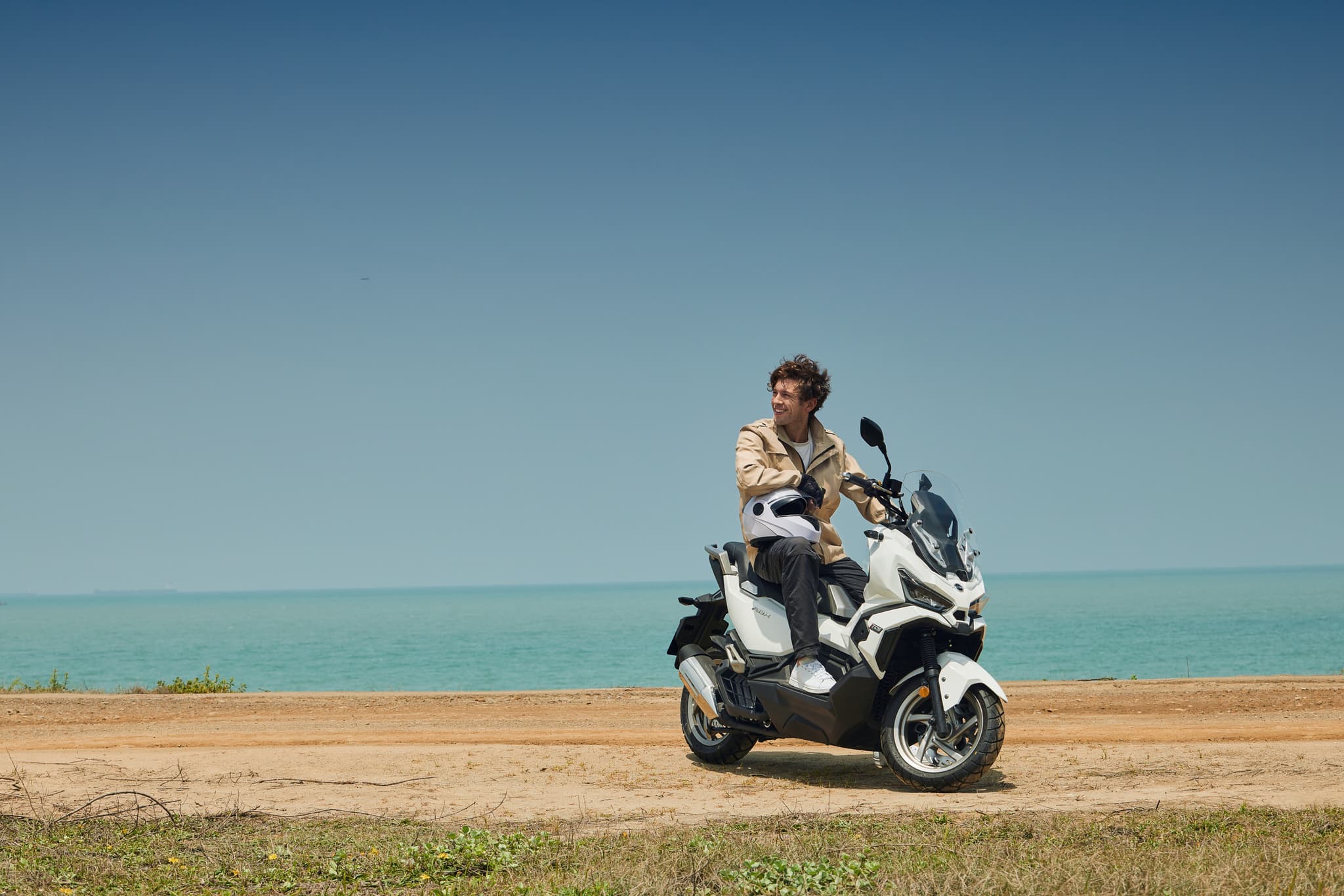Hombre en scooter SYM ADX 125 ABS TCS blanca con casco junto al mar